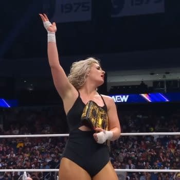 Toni Storm, wearing a black wrestling outfit, celebrates her victory in the ring, holding a championship belt above her head with her right hand raised. The crowd in the background is cheering her on.