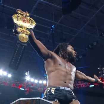 Trick Williams, celebrating his victory, holds the TNA Championship high above his head in a wrestling ring, with bright lights and a cheering crowd in the background.