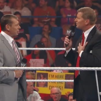 Vince McMahon, in a gray suit, stands on a wrestling ring with John Laurinaitis, who is wearing a black suit and using crutches. Fans holding colorful signs are visible in the background.