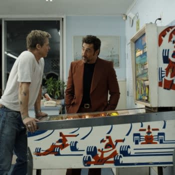 F1: Two men converse in a retro diner setting, leaning over a pinball machine featuring a colorful racing car design. One man is dressed casually in a gray t-shirt and jeans, while the other wears a stylish brown suit.