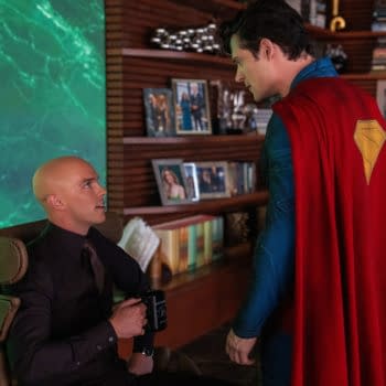 A scene featuring Superman, wearing a bright red cape with a yellow emblem on his back, standing with a bald man in a dark shirt who is seated and holding a coffee mug. The background includes a green patterned wall and several framed photographs on shelves.