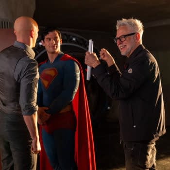 A behind-the-scenes moment featuring two men conversing, one dressed as Superman in a blue suit with a red cape, and a third man smiling and holding a script, likely a director on set.