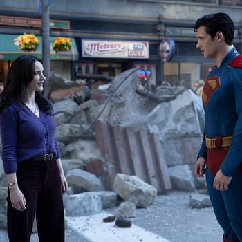 A scene featuring a woman in a purple sweater and a superhero in a blue suit with a red cape, standing amidst rubble in an urban setting. The background includes a coffee shop and hints of a crowd, suggesting a post-disaster scenario.