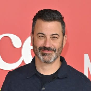 A man with dark hair and a beard smiles warmly against a vibrant red background featuring the word 'Netflix' in white letters. He is wearing a blue collared shirt.