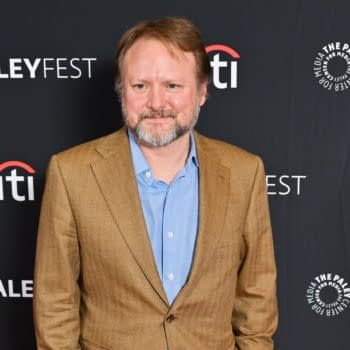Rian Johnson, a film director, stands smiling at a red carpet event, wearing a brown suit and blue shirt, against a black backdrop featuring the PaleyFest logo.