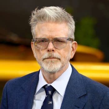 American director Christopher McQuarrie is wearing glasses and a blue suit, with a serious expression, at an event in London. The background includes hints of green foliage and a yellow element.
