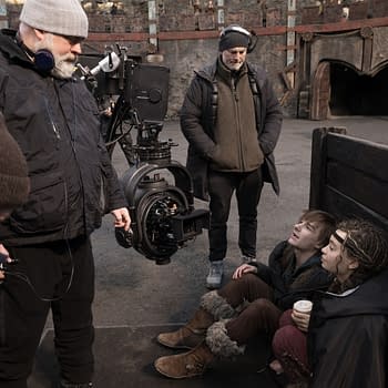 A behind-the-scenes moment from a film set featuring a director in a black jacket with headphones, discussing with two young actors seated on the ground. A large camera setup is visible, along with other crew members in warm clothing.