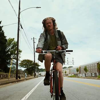 A scene from the film 'BUGONIA' (2025) featuring a man with long hair wearing headphones, riding a bicycle along a city street. He appears focused and is dressed in casual attire with a backpack.