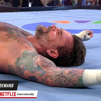 A defeated wrestler lies on the wrestling ring floor, showcasing tattoos on his arms and a weary expression. It is CM Punk in the aftermath of his match with John Cena at WWE Night of Champions in Saudi Arabia.