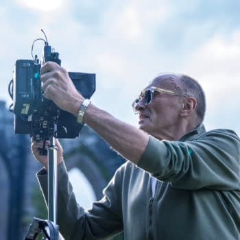 A photographer, wearing sunglasses, is adjusting a camera on a tripod in front of a historic stone archway under a bright sky.
