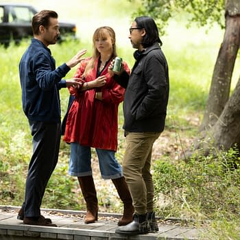 Three people engaged in conversation outdoors, surrounded by greenery; one person wears a red patterned top and brown knee-high boots, while another is in a navy jacket, and the third wears a black jacket. A black car is visible in the background.