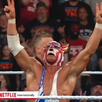 El Grande Americano, wearing a colorful mask and outfit, celebrates a victory in the wrestling ring, raising his arms in triumph. The excited crowd is visible in the background, reacting to the moment.