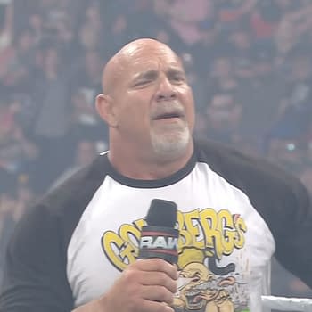 A muscular man in a 'Goldberg' t-shirt stands in a wrestling ring, holding a microphone. The crowd is visible in the background, cheering enthusiastically.