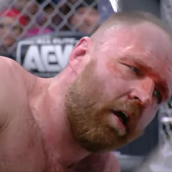 Jon Moxley shows a pained expression after winning a steel cage match against Kenny Omega, with visible wounds on his face and a backdrop of the cage.