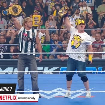 Logan Paul and John Cena stand in the WWE ring, both holding championship belts aloft, surrounded by an enthusiastic crowd. They each wear branded apparel, with Cena in a signature hat and shirt.