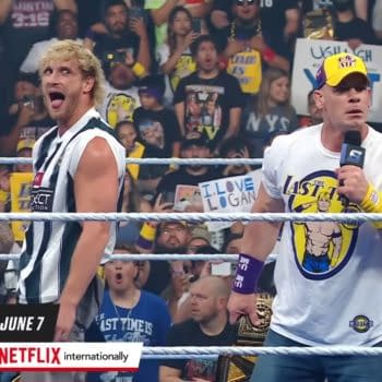 Logan Paul, wearing a black and white striped referee shirt, makes a playful expression while standing beside John Cena, who is wearing a white t-shirt and baseball cap, as they engage with an excited crowd at a WWE event. Fans can be seen in the background holding signs and cheering.