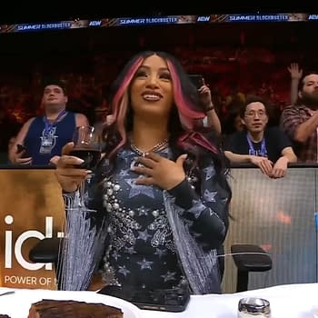 Mercedes Moné sits at a table enjoying a steak dinner during the AEW Summer Blockbuster event, holding a glass of drink and smiling, while fans are seen applauding in the background. A vase of flowers and a plate of food are on the table in front of her.