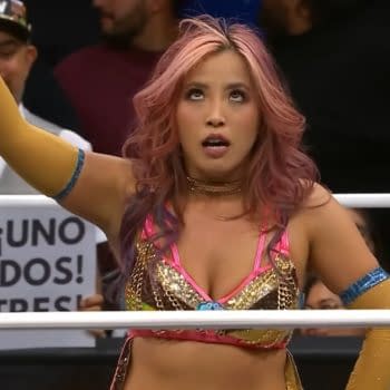 Mina Shirakawa, wearing a sparkling pink wrestling outfit with golden accents, raises her arm in victory after a win on AEW Collision, surrounded by cheering fans.