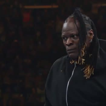 R-Truth, a wrestler with long braided hair, wearing a black hooded sweatshirt, stands in the center of a wrestling ring, with a tense expression on his face, as the crowd is visibly engaged in the background.