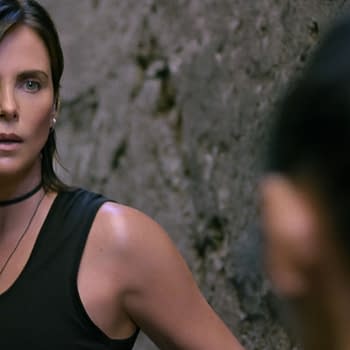 A scene from 'The Old Guard 2' features Charlize Theron as Andy, looking intently in a dramatic setting with walls in the background. The character appears focused and poised, conveying a sense of urgency.