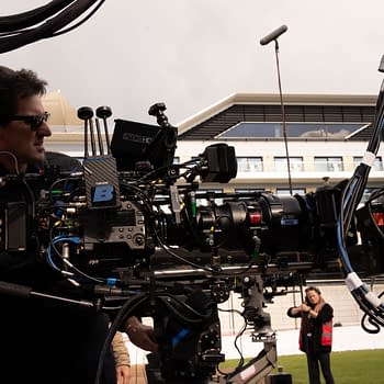 A film crew member operates a professional camera setup on a film set, with various cables and equipment visible, while another crew member prepares in the background. The setting appears to be an outdoor venue, suggesting a dynamic production environment.