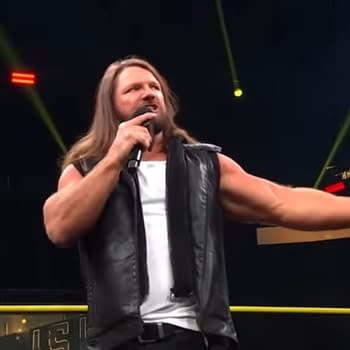 AJ Styles stands on stage at TNA Slammiversary, speaking into a microphone and gesturing enthusiastically toward the audience, wearing a black leather vest and tank top.