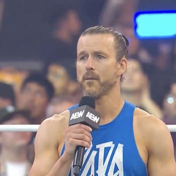 A wrestler, Adam Cole, stands in the ring addressing an audience, holding a microphone. He appears emotional while speaking to the crowd at AEW All In in Texas.