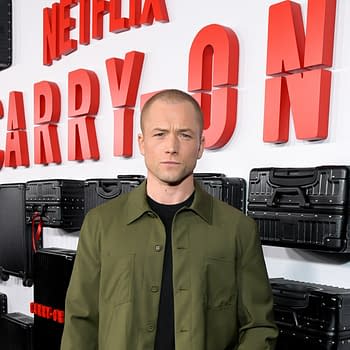 A man stands in front of a backdrop featuring the words "Netflix CARRY ON" in bold red letters, surrounded by black luggage cases. He is wearing an olive green jacket over a black shirt, displaying a serious expression.