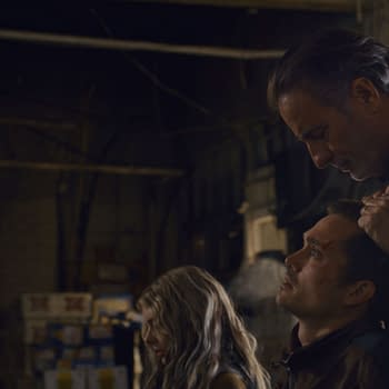 A tense scene from the film 'Eenie Meanie' featuring Samara Weaving as Edie, Karl Glusman as John, and Andy Garcia as Nico in a dimly lit industrial setting. The characters display intense emotions, with Nico holding John's head, suggesting a confrontation.
