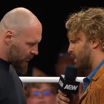 Jon Moxley and Hangman Adam Page stand face to face in a wrestling ring, both engaged in an intense exchange, with Moxley in a black shirt and Page in a brown fringed jacket.