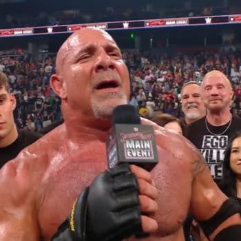 A muscular man in wrestling gear, holding a microphone, addresses a large crowd at an event. He shows signs of emotion, with sweat visible on his body, as he stands surrounded by supporters and fans.