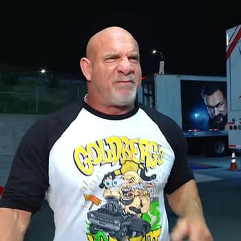 Goldberg is seen expressing determination while wearing a graphic t-shirt featuring his name and a cartoonish design. The background includes a WWE truck and a red classic car in the parking lot of WWE Raw.