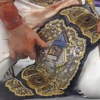 A close-up of a person's hand, belonging to Hangman Adam Page, reaching for a shiny AEW World Championship belt on the ground, adorned with jewels and intricate designs. The person's attire appears stained, suggesting an intense match.