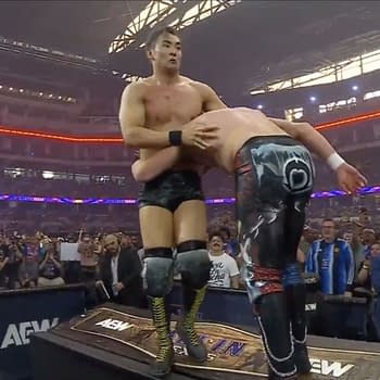Kazuchika Okada and Kenny Omega engage in a grappling contest on the announce desk during AEW All In Texas, with an energetic crowd visible in the background.