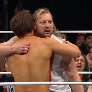 Kenny Omega and Kita Ibushi share an emotional embrace in the wrestling ring, surrounded by jubilant fans celebrating a milestone event. The two athletes display strong emotions as they momentarily set aside the intensity of competition.