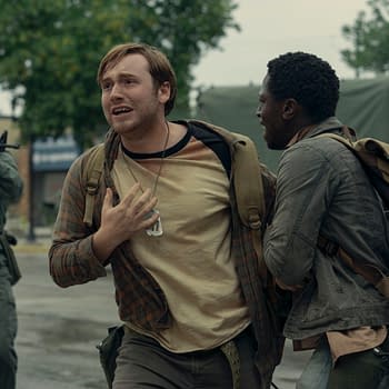 A tense moment from 'The Long Walk,' featuring two characters: one with a distressed expression and a plaid shirt, and another looking concerned, both wearing backpacks. In the background, a soldier is aiming a firearm, adding to the scene's intense atmosphere.