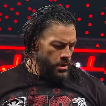 A focused Roman Reigns stands in the wrestling ring on WWE Raw, wearing a black shirt. His hair is long and wet, and there is a backdrop of bright red lights illuminating the scene.