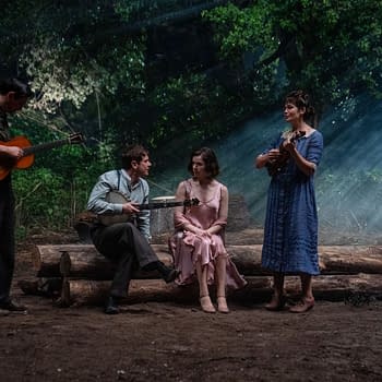 A scene featuring four performers in a forest setting, with two holding string instruments and two seated, creating a cozy atmosphere illuminated by soft, natural light filtering through the trees.