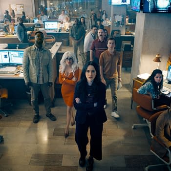 A scene from the film 'Superman' featuring a group of diverse characters in an office setting, with several people gathered around computer desks, showing expressions of concern and focus as they watch something on screens.