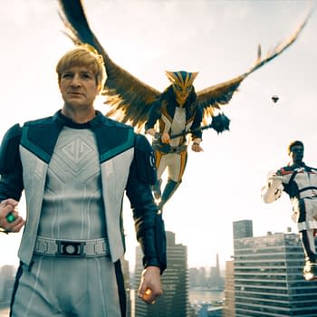 A scene from a superhero film featuring a main character in a futuristic outfit with a glowing green object, flanked by two flying characters, one with bird-like wings and the other seated in a high-tech chair, set against a city skyline.