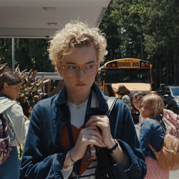 A scene outside a school showing a young woman with curly hair and glasses, looking somber while holding a bag. The background features students and a yellow school bus, with a memorial display reading "LOOK STRONG".