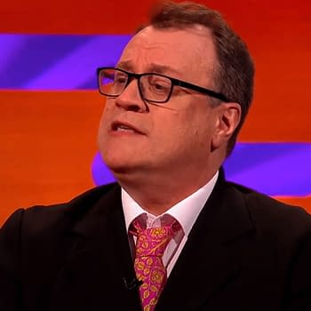 Russell T. Davies in a dark suit speaks passionately, wearing a colorful pink and orange patterned tie, against a vibrant orange and purple backdrop.