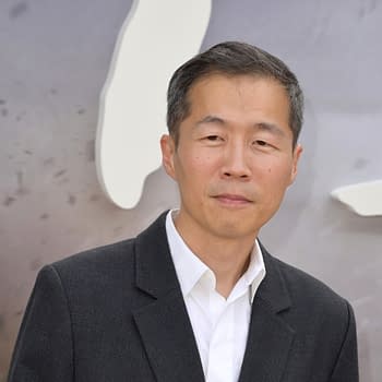 A man with short black hair is wearing a gray blazer over a white shirt, standing against a blurred background. He has a neutral expression and appears poised.