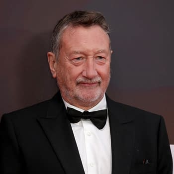 A half-length portrait of a middle-aged man with a greying beard, wearing a black tuxedo, and smiling while attending a film event. The background features a blurred dark gradient, enhancing the focus on his formal attire and expression.