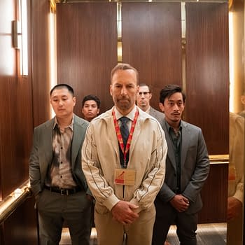 A group of five men stands solemnly in an elevator, dressed in suits and casual attire, with one man prominently wearing a badge around his neck. The setting features polished wooden walls and soft lighting, creating a tense atmosphere.
