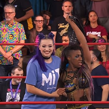 Two female wrestlers celebrate a victory in a wrestling ring. One wrestler, wearing a blue shirt, smiles broadly while the other, dressed in a colorful outfit, raises a championship belt triumphantly.