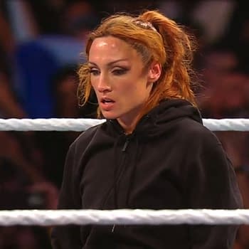 Becky Lynch standing in a wrestling ring, visibly focused and surveying the arena, wearing a black hoodie. The backdrop features a cheering crowd.