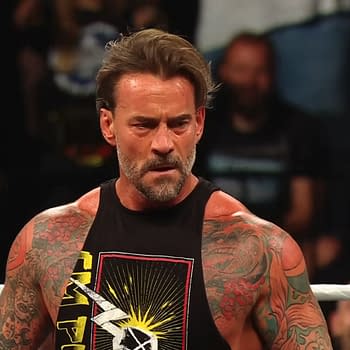 A muscular man with tattoos, CM Punk, appears on WWE Raw, wearing a black tank top with a graphic design. He looks intense, engaging with the audience.