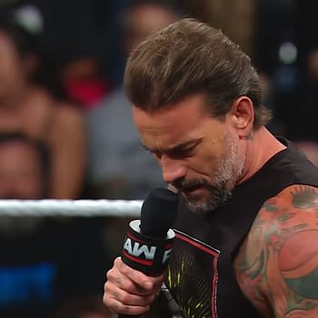 A professional wrestler, CM Punk, holds a microphone while appearing thoughtful during a segment on WWE Raw. He's wearing a sleeveless shirt and has multiple tattoos visible on his arms.