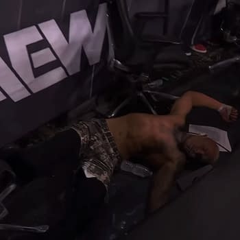 Darby Allin lies on the ground amid debris from the announce table, suggesting he has just been thrown out of the wrestling ring by Claudio Castagnoli during a match.